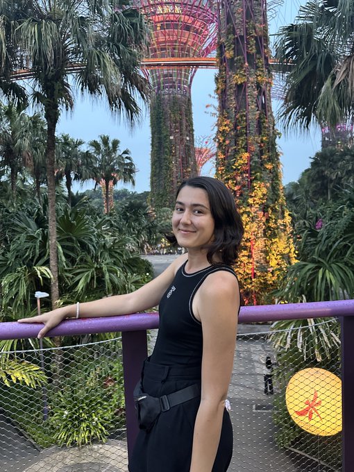 A young woman poses in front of the Supertree Grove at Gardens by the Bay, featuring the iconic flower-dome structures with vibrant purple lighting after watching the Garden Rhapsody show on her first day in Singapore.
