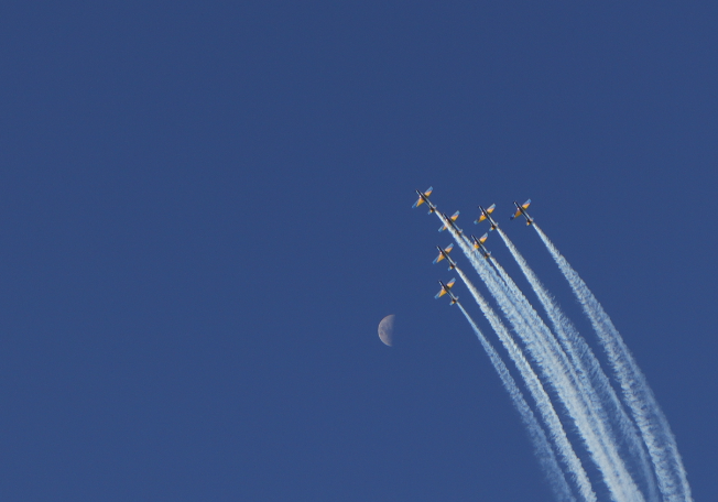 A formation of jets flies in close proximity across a clear blue sky, leaving white contrails behind them, with a crescent moon visible in the background.