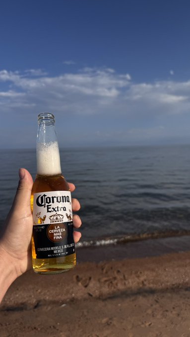 A hand cheers a Corona Extra beer bottle on a sunny beach, matching the casual "gm!" (good morning) vibe with ocean waves and blue skies in the background.