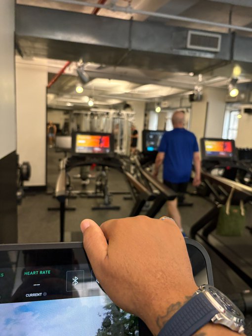 A first-person view from a treadmill in a gym shows the photographer's hand resting on the console, while a man in a blue shirt walks ahead on an adjacent machine.