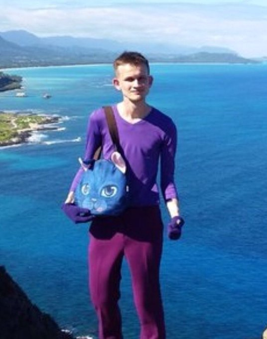 A young man smiling while standing on a rocky coastal cliff overlooking a blue ocean, wearing a purple shirt and maroon pants, and carrying a blue cat-shaped crossbody bag.
