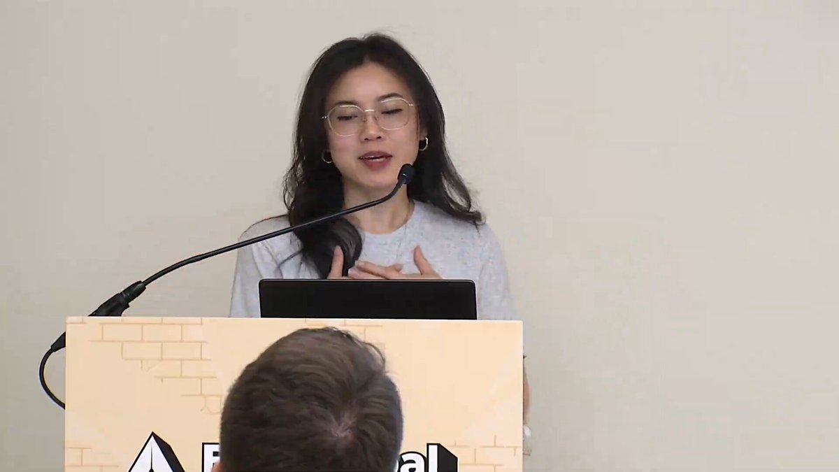 The image features a woman standing behind a podium, giving a speech or presentation. She appears to be wearing glasses as she speaks into a microphone connected to the podium. The setting seems to be indoors, with the room featuring a gray wall on one side of the frame and a brick wall on another side. In front of her is an object resembling a book, which could potentially be part of her presentation or speaking material.