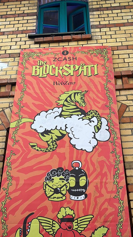 The image features a large, colorful banner hanging from a brick building with a window. The banner displays an illustration of a zebra and a unicorn, along with some text that reads "Algorand $ALGO". The banner appears to be advertising or promoting something related to Algorand, possibly a product or service.