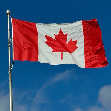 The Canadian flag, red and white with the central maple leaf, waving on a flagpole against a blue sky with scattered clouds.