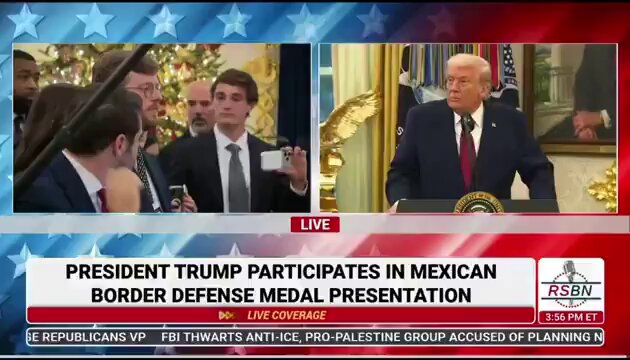 The image shows a news broadcast with two men sitting at a table, one of whom appears to be President Trump. The other man is holding up a microphone, suggesting that they are discussing or presenting something related to the President's presidency. The background features an American flag and stars, indicating that this event might be taking place in the United States.