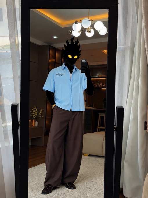 A person takes a mirror selfie wearing a light blue short-sleeved button-up shirt, brown wide-leg pants, and black shoes, with their face obscured by a black flame graphic and glowing yellow eyes.