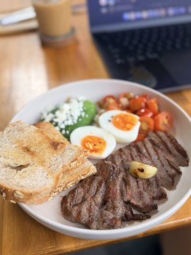 A hearty breakfast spread featuring grilled steak with herb butter, a soft-boiled egg over greens with feta, cherry tomatoes, and crusty bread, enjoyed alongside an iced coffee and laptop—setting the scene for a productive morning the poster is greeting with a playful "Good morning xaggots."