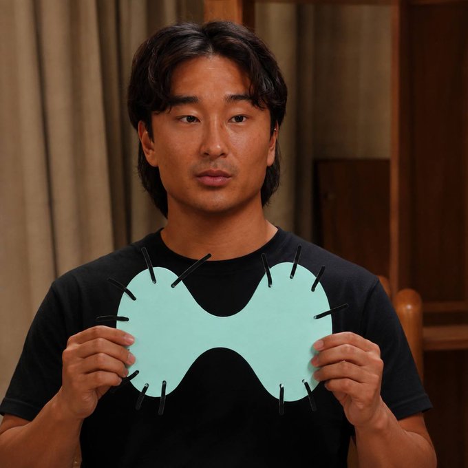Actor Simu Liu holding a light blue, mask-like prop with protruding black sticks in front of his chest.