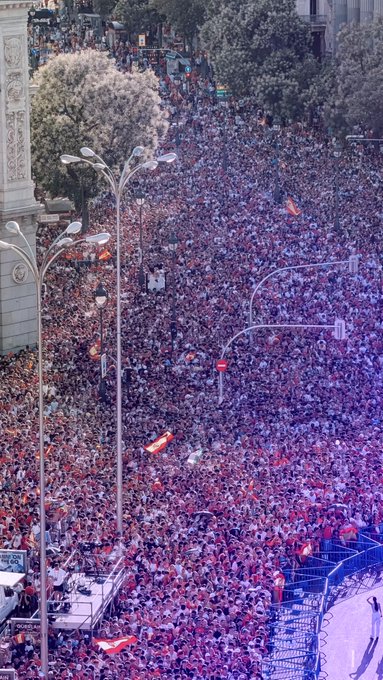 A massive crowd of fans fills the streets of Madrid, celebrating the Spanish national team's World Cup championship victory parade, with people waving Spanish flags and packing every inch of the avenue. The sea of supporters, stretching as far as the eye can see, reflects the immense excitement described in the post about Spain's triumphant return home.
