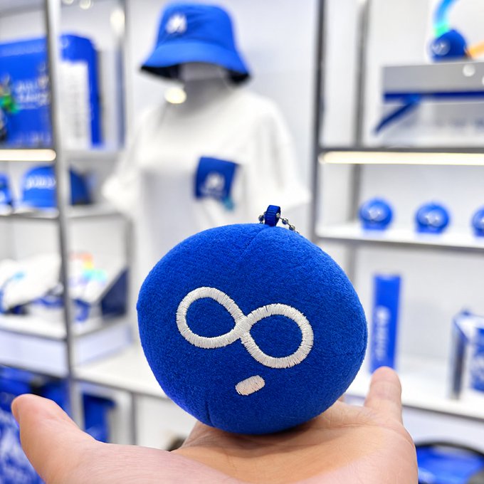 A hand holds up a round blue plush mascot with white glasses design, photographed inside what appears to be a MEXC merchandise booth featuring branded apparel, t-shirts, and display shelves in the blurred background. The plush serves as a promotional giveaway item tied to the MEXC crypto exchange's TOKEN2049 social media contest.