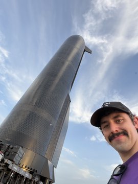 A man with a mustache wearing a black cap and purple shirt takes a selfie in front of a large metallic rocket or tower against a blue sky.