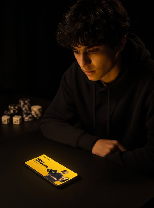 A young man in a black hoodie stares intently at his glowing smartphone in a dimly lit room, with scattered dice on the table beside him—visually framing the tension between gambling instinct and the disciplined, ETF-driven investing strategy highlighted in the accompanying post about Gen Z's surprisingly measured behavior on Binance.