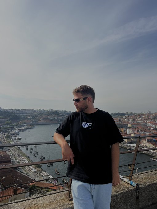 A man in a black Vans t-shirt and sunglasses leans against a railing overlooking the Douro River and historic cityscape of Porto, Portugal, with his tag "@coldtrz" visible in the background, matching the caption's request to drop a $SOL address as he poses in a scenic travel-style photo.