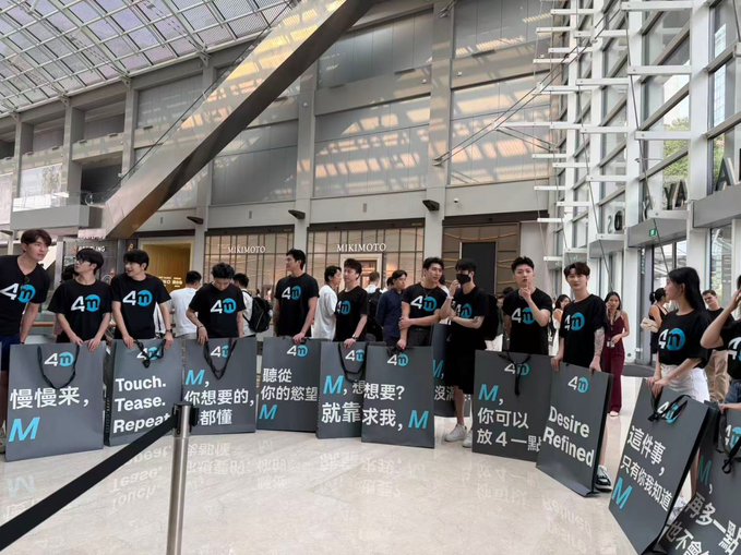 A group of people wearing black shirts with the number '4D' stands in a modern glass-walled atrium, displaying signs with Chinese and English phrases such as 'Touch. Tease. Repeat' and 'Desire Refined.'