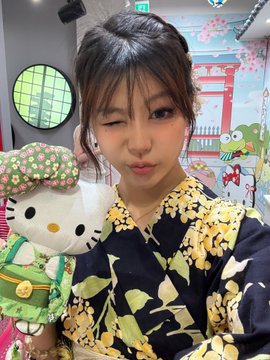 A selfie of a woman in a floral kimono making a playful pout, holding a Hello Kitty plush wearing a green headscarf and apron.