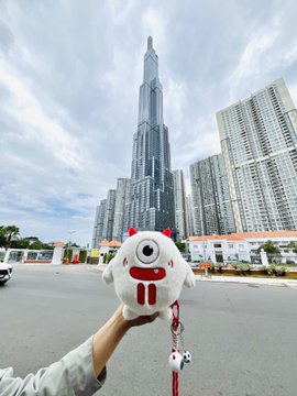 Hand holding a small mascot plush toy in front of the Landmark 81 skyscraper in Ho Chi Minh City, Vietnam, with surrounding high-rise buildings.