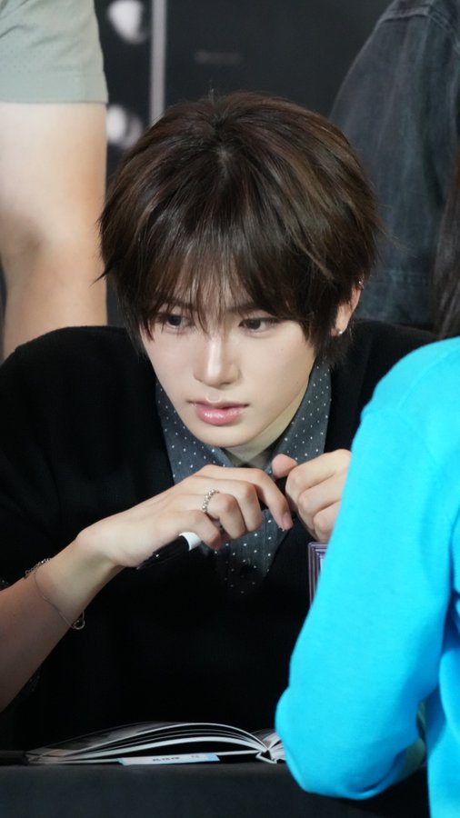 A young man with brown hair wearing a black vest and a polka-dot shirt looks down at an open notebook at what appears to be a fan signing event.