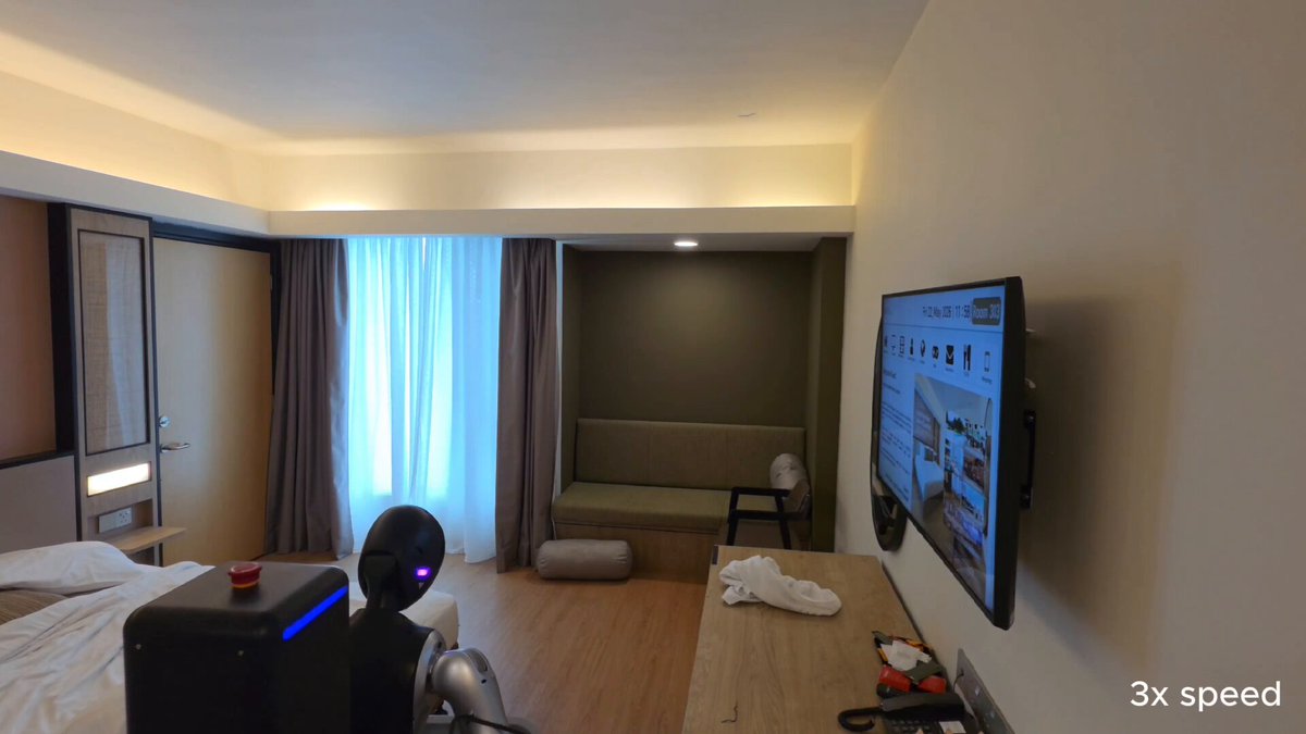 The image shows a room that appears to be a hotel room, possibly a motel or a suite. The room has a flat-screen TV mounted on the wall above a couch. A bed can also be seen in the room. There are two people present in the scene - one person is sitting on the couch and another person is standing near the TV. 

The room features a window with curtains, allowing natural light to enter the space. The overall atmosphere of the room seems comfortable and inviting, making it an ideal place for relaxation or entertainment.