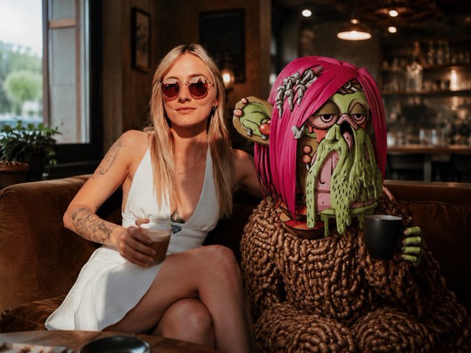 Photograph of a blonde woman in a white dress and red sunglasses sitting on a brown leather couch next to a Cthulhu-like creature with pink hair and a tentacled face, both holding coffee mugs in a dimly lit cafe.