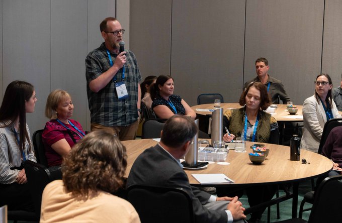 A photo of a conference or networking event where a man with glasses and a plaid shirt is speaking into a microphone while seated at a round table with several attendees.