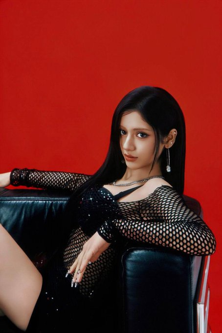 A glamorous portrait of a woman with long dark hair, wearing a black mesh dress over a dark outfit, seated against a vibrant red backdrop.