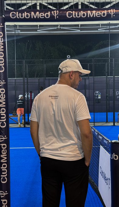 A man wearing a white OZNAO branded t-shirt and white cap standing inside a blue padel tennis court branded with "Club Med" logos at night.