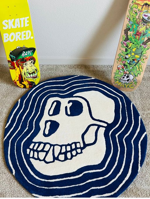 The image features a blue rug with a white skull design, placed on a gray carpeted floor. Next to the rug are two skateboards - one yellow and one brown. The background of the image includes a wall painted white, which contrasts with the blue rug and adds depth to the scene. The text "Skate bored" is visible in the image as well.
