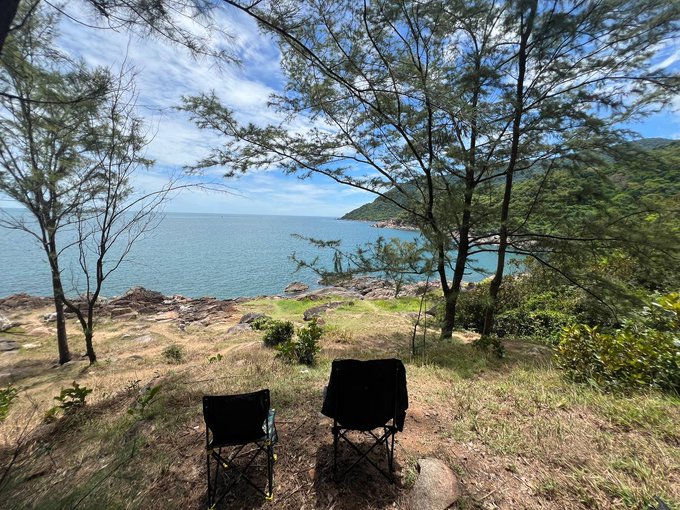 Two folding camping chairs set up on a grassy hill overlooking a scenic view of the sea, rocky shoreline, and forested mountains under a blue sky.