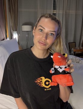 A blonde woman wearing a black 'BC CAMP' t-shirt, holding a red Angry Birds plush toy, seated in what appears to be a hotel room.