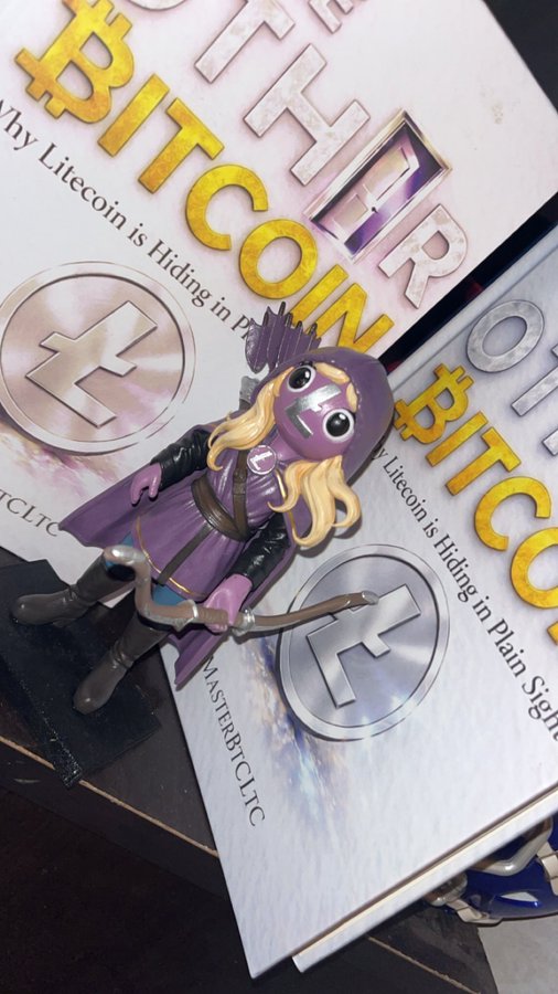 A small figurine of a purple-clad archer with a bow, positioned on top of books titled 'The Tip of Bitcoin' and 'Litecoin is Hiding in Plain Sight' by 'MasterBTC LTC'.