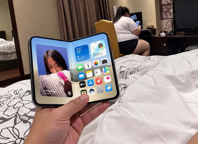 A first-person view of a hand holding an unfolded foldable smartphone, likely a Samsung Galaxy Z Fold, displaying the home screen with widgets and apps, with a bed and another person using a phone in the background of a hotel room.