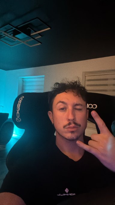 A man sits in a black Noblechairs gaming chair (logo visible on the headrest) flashing a peace sign and winking at the camera in a room lit by ambient blue lighting, setting the mood for his Assetto Corsa racing simulator session.