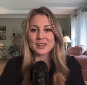 A blonde woman speaking into a microphone in a living room setting with a couch, fireplace, and lamps visible in the background.