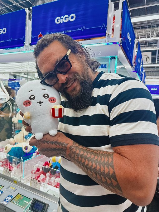 A bearded man with tattoos and dark sunglasses smiles while holding a Chiikawa plush toy in front of a 'GiGO' arcade game machine.