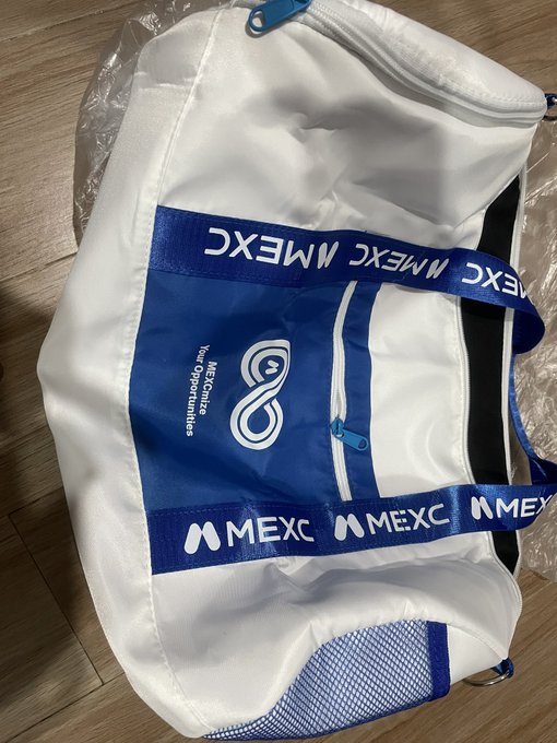 A white promotional drawstring bag with blue trim and 'MEXC' branding featuring the MEXC logo and 'Your opportunities' tagline, partially visible in plastic packaging.