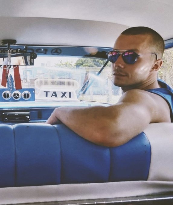 A man wearing a tank top and reflective sunglasses sits in the back of a taxi, looking over his shoulder toward the camera.
