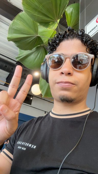 A selfie of a young man wearing sunglasses and black headphones, making a peace sign with his hand in front of a large green fan palm plant.