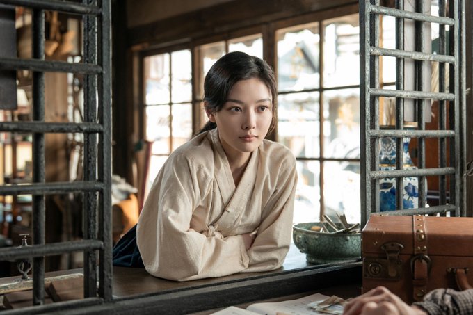 A young woman in a traditional beige hanbok leans pensively at a wooden window frame, her contemplative gaze framed by weathered iron lattice. The scene's vintage Korean setting—with its old suitcase, ceramic bowl, and rustic interior—evokes the period drama atmosphere of "100 Days of Deception," hinting at the story's tension just one day before the drama's premiere.