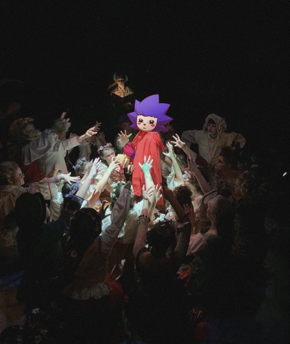 A theatrical performance scene featuring a person in a red costume with a spiky purple and blue character headpiece being lifted by a crowd of performers with raised hands on a dark stage.