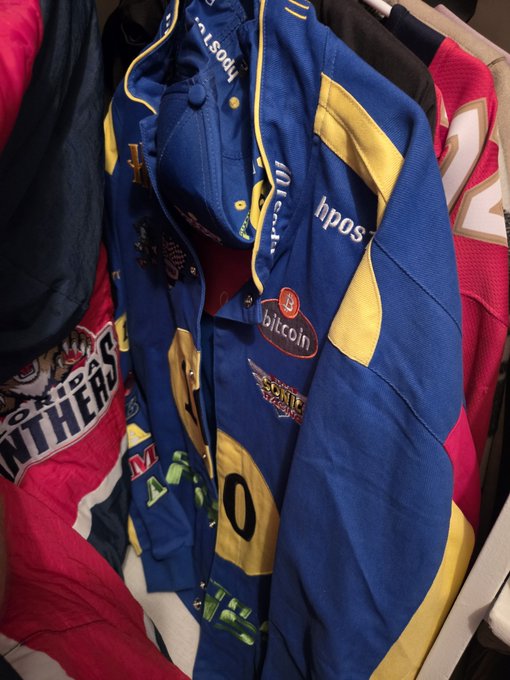 A blue and yellow varsity-style jacket with "hpos" branding and a "bitcoin" patch sits among other clothing items, with a matching baseball cap resting on top. The jacket features sponsor-style lettering and additional patches, matching the description of a collectible HPOS10I item being sold.
