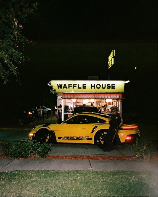 A nighttime photo of a yellow Porsche 911 GT3 RS parked in front of a brightly lit Waffle House restaurant, with a person wearing sunglasses leaning against the car.