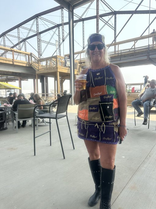 A blonde woman wearing sunglasses, a captain's hat, and a colorful patchwork Crown Royal-patterned dress stands holding a beer on an outdoor patio at what appears to be a beachside venue.