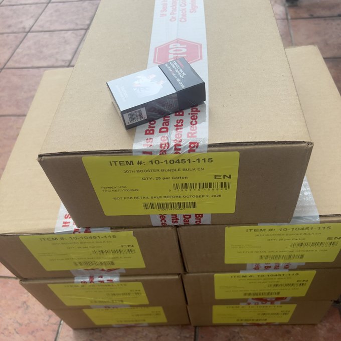 Several cardboard boxes are stacked on a tiled floor, labeled 'ITEM #: 10-10451-115', '30TH BOOSTER BUNDLE BULK EN', with 'NOT FOR RETAIL SALE BEFORE OCTOBER 2, 2026' printed on them.