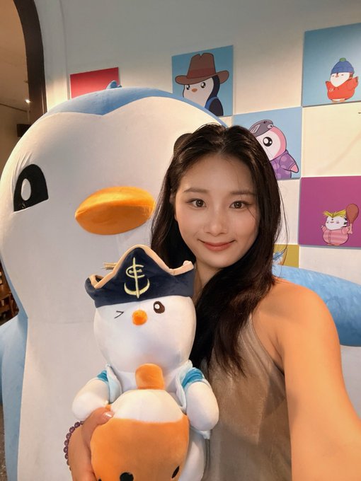 A young woman smiles warmly while holding a small plush penguin toy dressed in a pirate hat with an anchor emblem, posing next to a large penguin mascot in a room decorated with colorful Pudgy Penguins artwork on the walls, fitting her cheerful "Good morning, Pudgy Penguins" greeting to the brand's Chinese community.