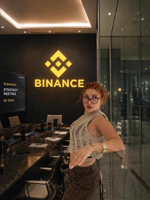 A woman with red hair and glasses poses in front of a large illuminated Binance logo, with a presentation screen displaying "Strategy Meeting Q2 2024" beside her in a modern conference room. The image visually reinforces the post's theme by capturing an on-site moment at Binance's headquarters during a strategic meeting, symbolizing the company's response to mounting regulatory pressures across Europe, the U.S., and Türkiye.