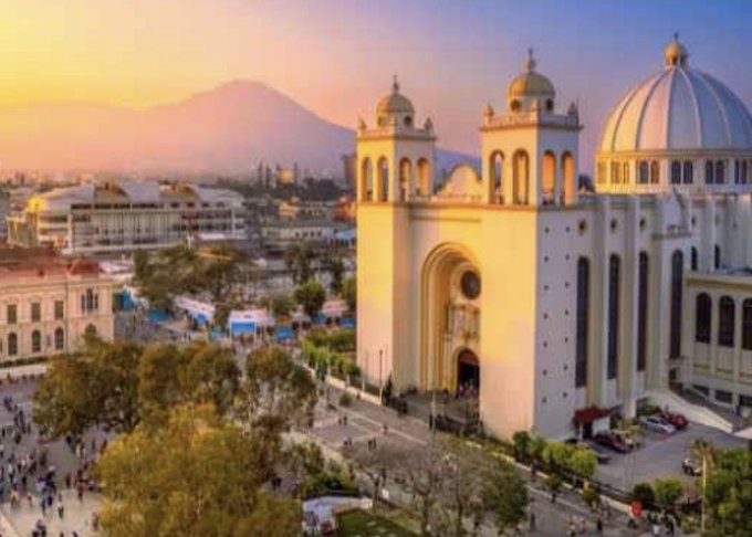 A stunning golden-hour view of the Basilica of Our Lady of Guadalupe in Mexico City, with the iconic Popocatépetl volcano visible in the hazy distance behind it. The post humorously refers to Mexico as "Bitcoin Country," playing on El Salvador's 2021 adoption of Bitcoin as legal tender, while suggesting October as a good time to visit this landmark destination.