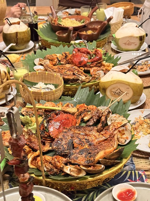 An elaborate seafood feast spread on a table features grilled crab, fish, shellfish, and rice, accompanied by fresh coconuts and various dipping sauces on banana leaves.