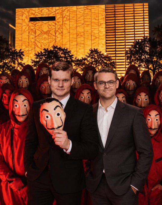 Two men in business suits stand in front of a large group wearing red hooded jumpsuits and Salvador Dalí masks, holding one of the masks in front of a golden building at dusk.