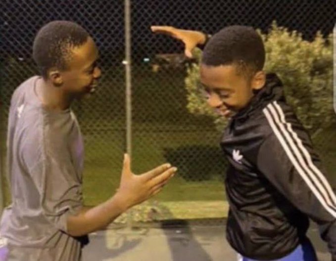 Two young men standing near a fence at night, with a hand visible in the background pointing at the head of the person on the right.