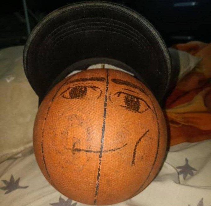 A close-up of a basketball with a hand-drawn cartoon face on it, wearing a black cylindrical object like a hat, resting on a patterned fabric.